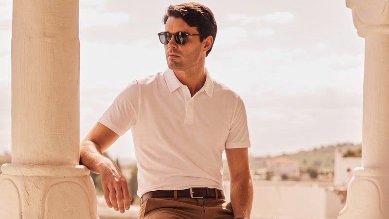 Man in white polo shirt and beige pants sitting on a stone wall with columns in the background.